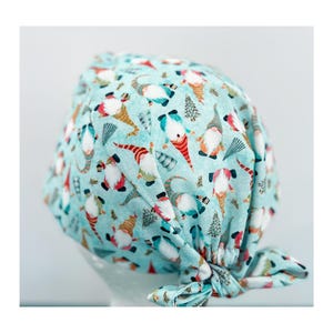 May include: A light blue surgical scrub cap with a repeating pattern of gnomes, Christmas trees, and other festive elements. The gnomes have white beards, red hats, and orange noses. The cap is tied in a knot at the back.