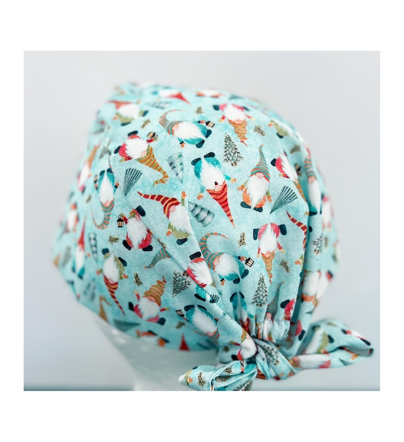 May include: A light blue surgical scrub cap with a repeating pattern of gnomes, Christmas trees, and other festive elements. The gnomes have white beards, red hats, and orange noses. The cap is tied in a knot at the back.