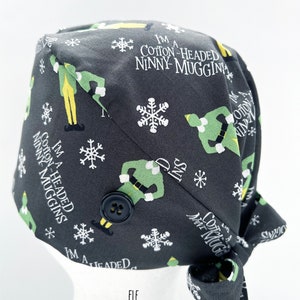 May include: A dark gray surgical scrub cap with a repeating pattern of cartoon elves, snowflakes, and the phrase "I'M A COTTON-HEADED NINNY MUGGINS." The cap has a black button and is tied at the back. The word "ELF" is printed at the bottom.