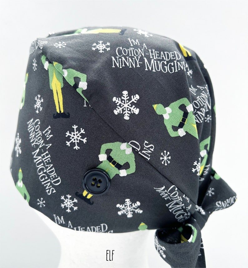 May include: A dark gray surgical scrub cap with a repeating pattern of cartoon elves, snowflakes, and the phrase "I'M A COTTON-HEADED NINNY MUGGINS." The cap has a black button and is tied at the back. The word "ELF" is printed at the bottom.