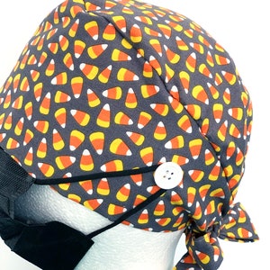 May include: A gray surgical scrub cap with a repeating pattern of candy corn in orange, yellow, and white. The cap has a white button and black elastic loops for attaching a face mask. A black face mask is attached.