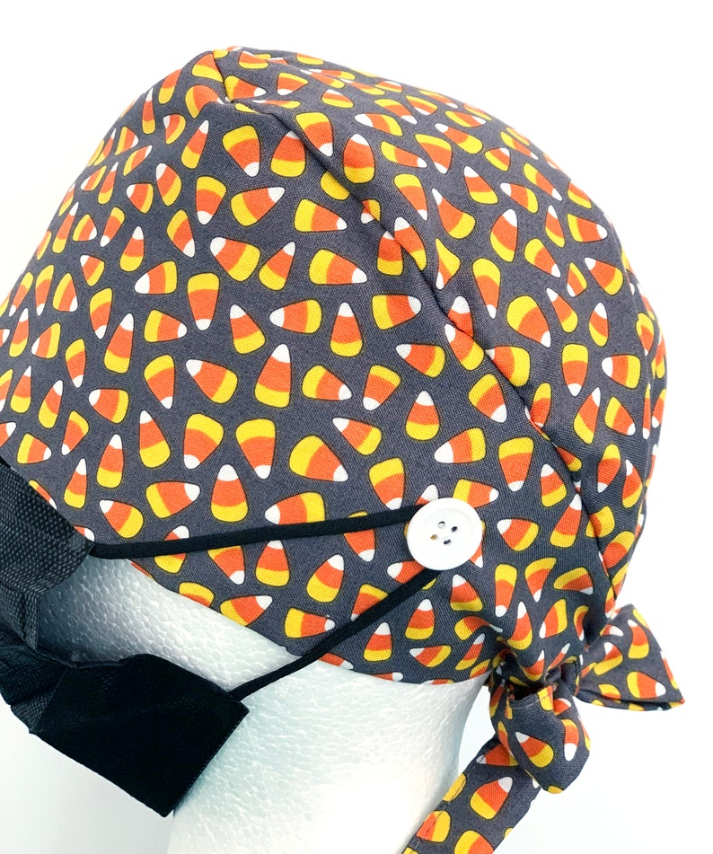 May include: A gray surgical scrub cap with a repeating pattern of candy corn in orange, yellow, and white. The cap has a white button and black elastic loops for attaching a face mask. A black face mask is attached.