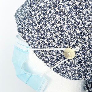 K&ouml;nnte beinhalten: Eine wei&szlig;e OP-M&uuml;tze mit einem blauen Blumenmuster und einem wei&szlig;en Knopf an der Seite. Die M&uuml;tze wird auf einem wei&szlig;en Mannequin-Kopf mit einer blauen chirurgischen Maske getragen, die an der Seite befestigt ist.