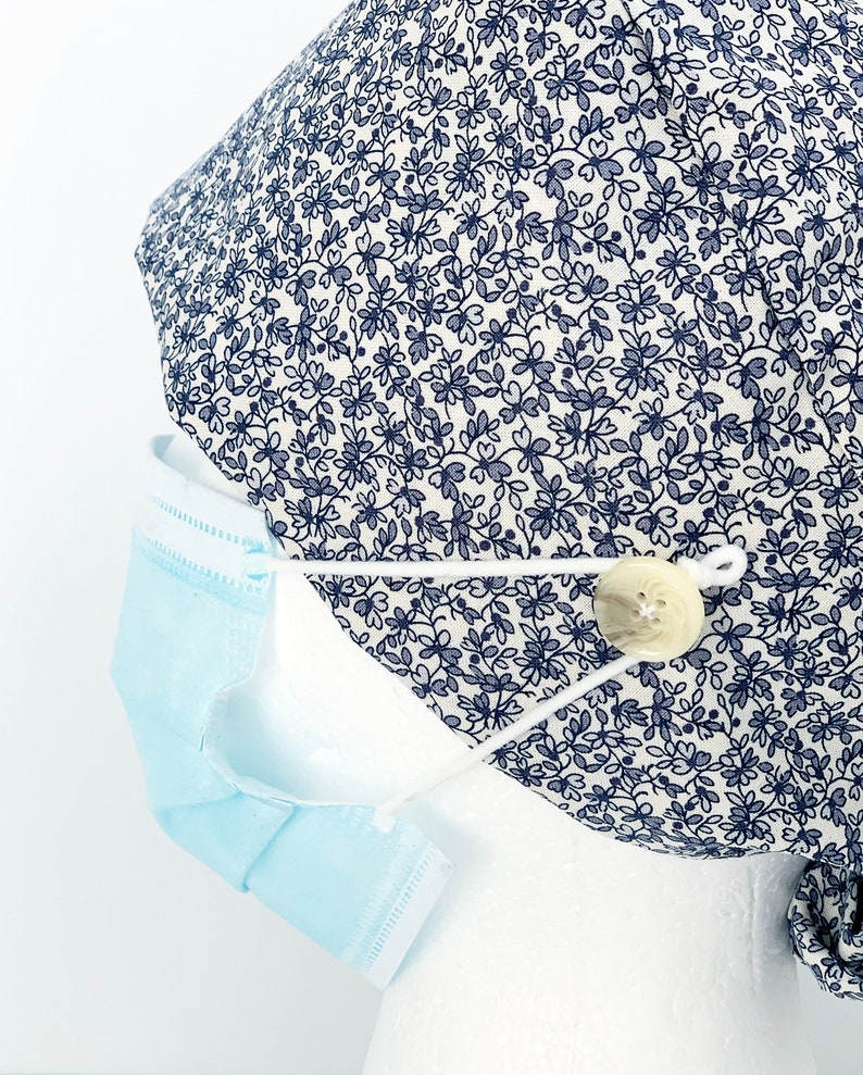 K&ouml;nnte beinhalten: Eine wei&szlig;e OP-M&uuml;tze mit einem blauen Blumenmuster und einem wei&szlig;en Knopf an der Seite. Die M&uuml;tze wird auf einem wei&szlig;en Mannequin-Kopf mit einer blauen chirurgischen Maske getragen, die an der Seite befestigt ist.