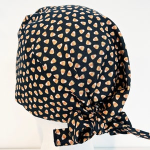 May include: A black scrub cap with a repeating pattern of candy corn in orange, yellow, and white. The cap is tied in a bow at the back. The text "CANDY CORN" is printed in a handwritten font at the bottom.