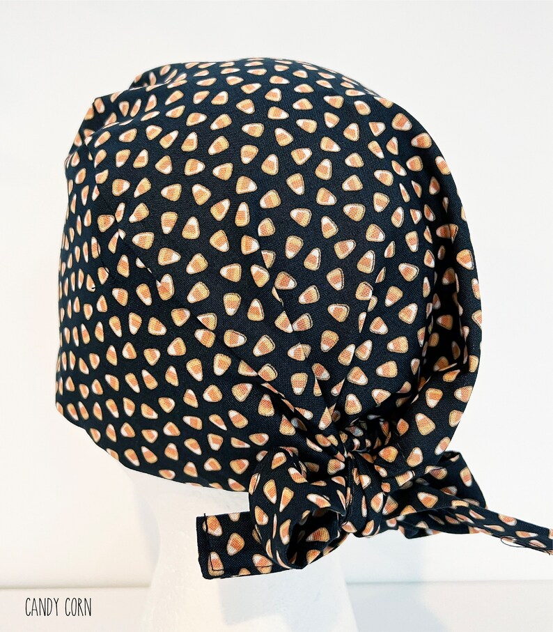 May include: A black scrub cap with a repeating pattern of candy corn in orange, yellow, and white. The cap is tied in a bow at the back. The text "CANDY CORN" is printed in a handwritten font at the bottom.