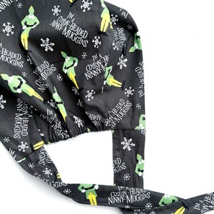 May include: A black surgical scrub cap with a repeating pattern of green elves, snowflakes, and the text "I'm a Cotton-Headed Ninny Muggins" in white. The cap has ties for a secure fit.