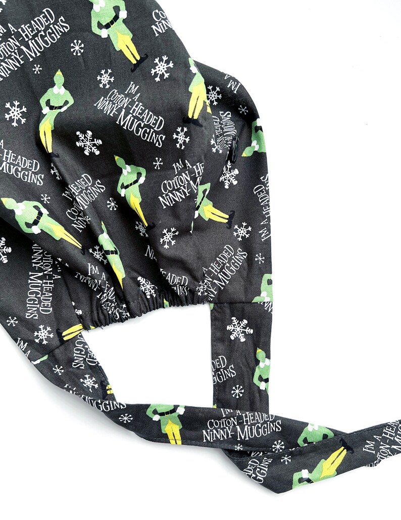 May include: A black surgical scrub cap with a repeating pattern of green elves, snowflakes, and the text "I'm a Cotton-Headed Ninny Muggins" in white. The cap has ties for a secure fit.