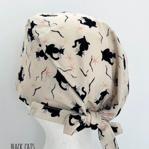 May include: Cream-colored surgical scrub cap with a repeating pattern of black cat silhouettes, starbursts, and curved lines. The cap has a tie closure and the text "BLACK CATS" is visible.