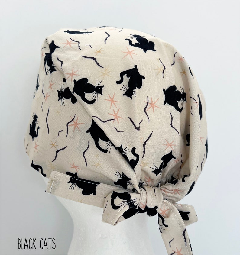 May include: Cream-colored surgical scrub cap with a repeating pattern of black cat silhouettes, starbursts, and curved lines. The cap has a tie closure and the text "BLACK CATS" is visible.