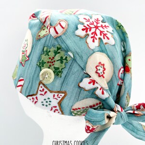 May include: A light blue surgical scrub cap featuring a pattern of Christmas cookies in red, white, and green. The cap has a button detail and a tie closure. The text "CHRISTMAS COOKIES" is printed at the bottom.