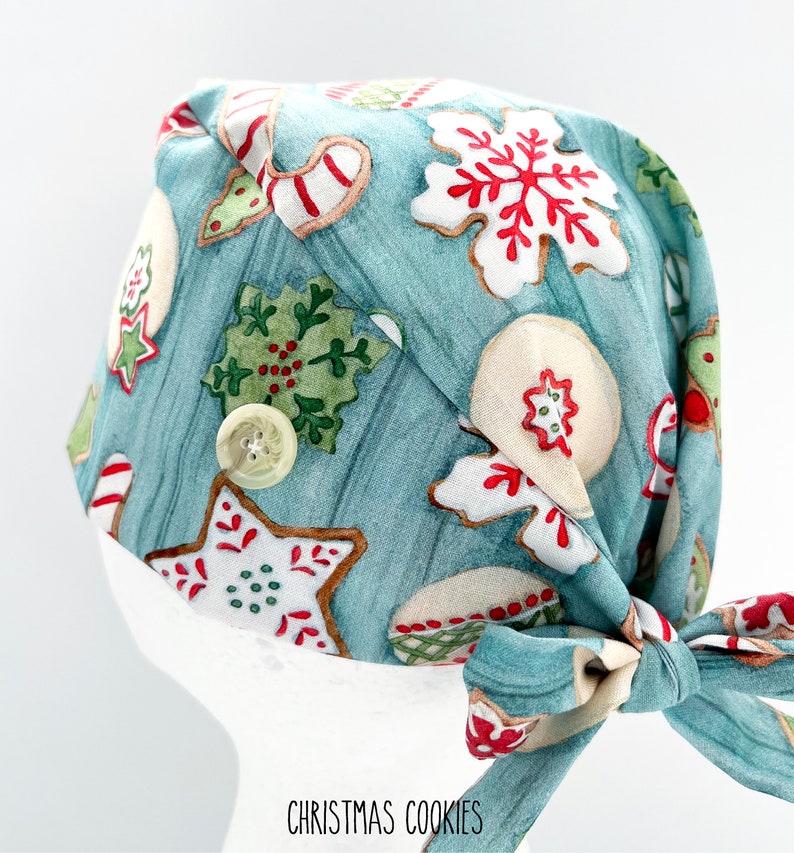 May include: A light blue surgical scrub cap featuring a pattern of Christmas cookies in red, white, and green. The cap has a button detail and a tie closure. The text "CHRISTMAS COOKIES" is printed at the bottom.