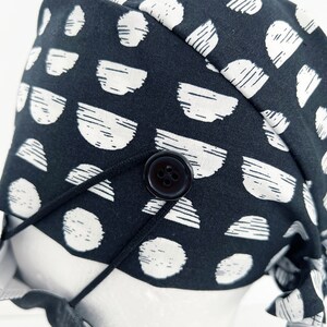 May include: Black and white patterned fabric scrub hat with a black button and adjustable tie strings.