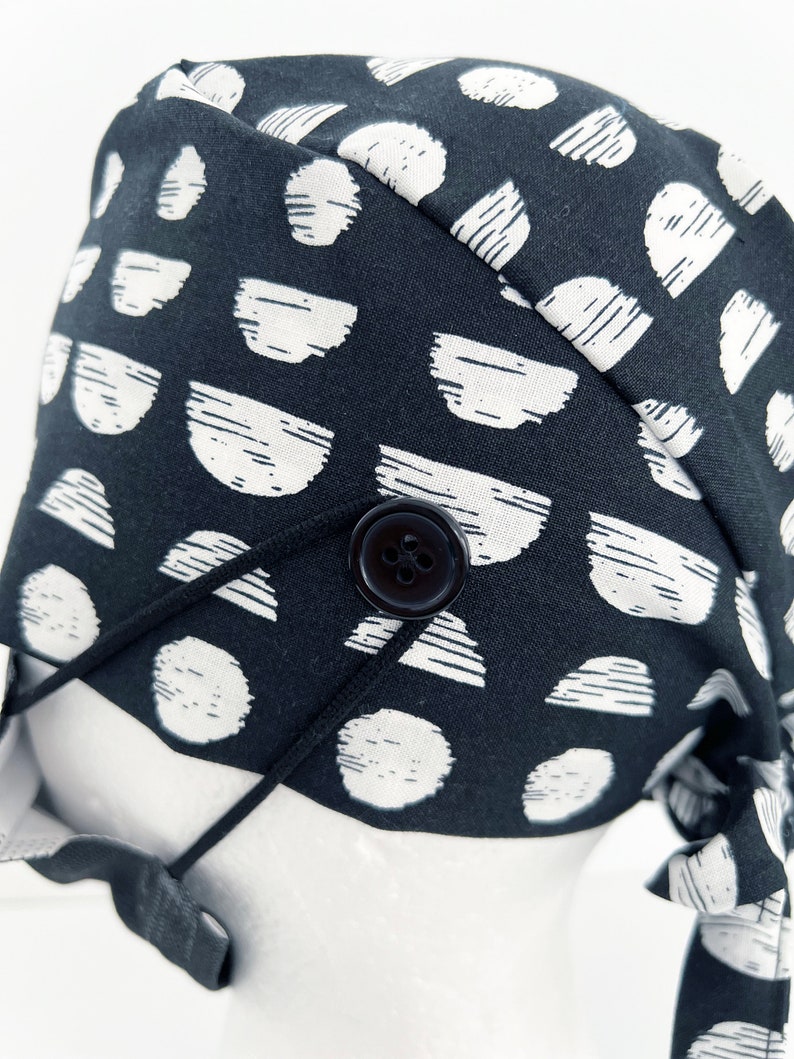 May include: Black and white patterned fabric scrub hat with a black button and adjustable tie strings.