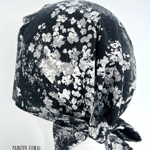 May include: Black and white patterned surgical scrub hat with a tie at the back. The pattern is a white floral design on a black background. "Painted Floral" is written in white text at the bottom of the image.