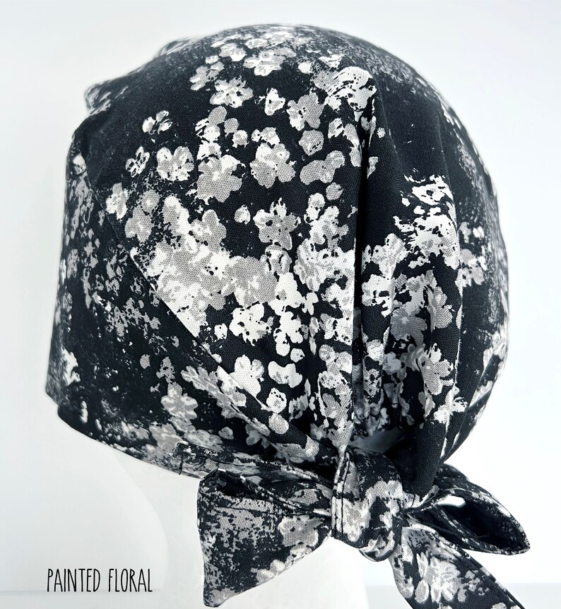 May include: Black and white patterned surgical scrub hat with a tie at the back. The pattern is a white floral design on a black background. "Painted Floral" is written in white text at the bottom of the image.