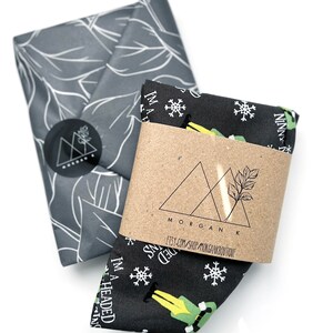 May include: Two folded fabric items. One is wrapped in gray paper with a white leaf pattern and a black sticker. The other is black with white snowflakes and a green elf graphic. A brown paper band is wrapped around the black fabric with the text "I'M A HEADED NINNY".