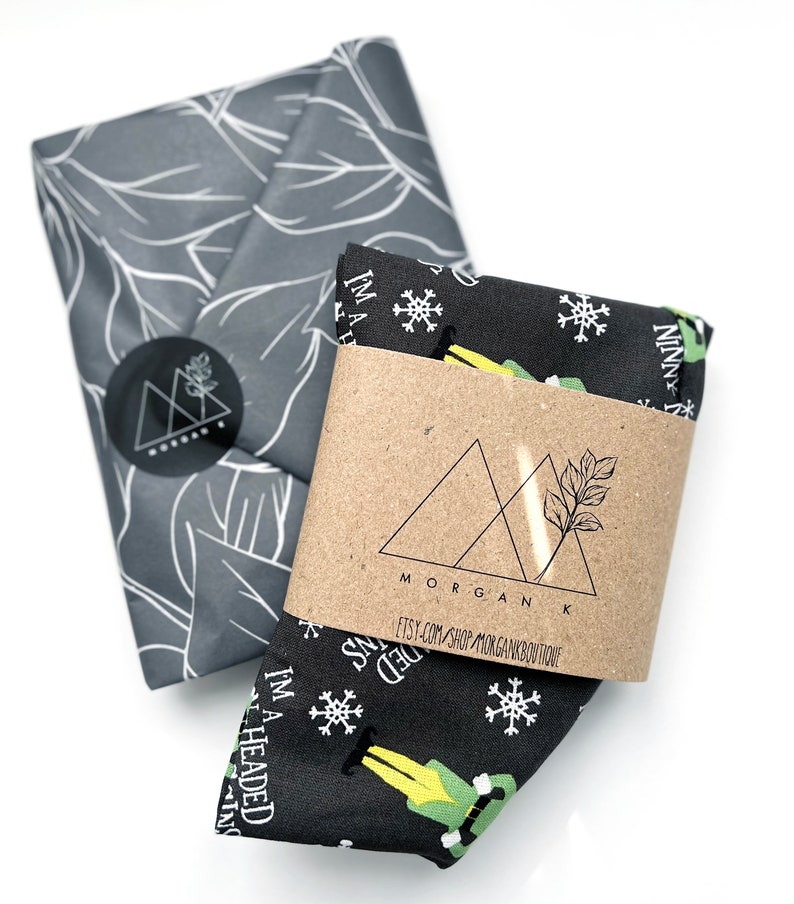 May include: Two folded fabric items. One is wrapped in gray paper with a white leaf pattern and a black sticker. The other is black with white snowflakes and a green elf graphic. A brown paper band is wrapped around the black fabric with the text "I'M A HEADED NINNY".