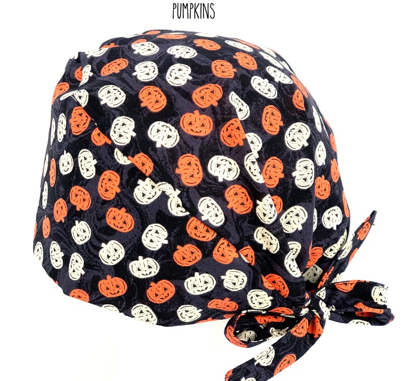May include: A black surgical scrub cap with a repeating pattern of orange and white jack-o'-lanterns. The cap is tied at the back with matching fabric. The word "PUMPKINS" is printed at the top.