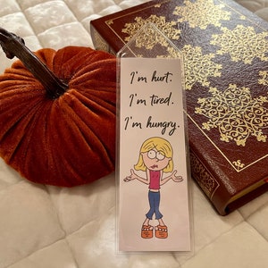 May include: A white bookmark with a cartoon illustration of a girl with blonde hair and a pink shirt. The bookmark has the text "I'm hurt. I'm tired. I'm hungry." written on it.