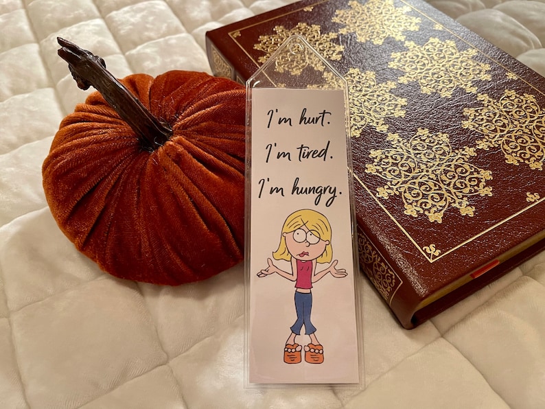 May include: A white bookmark with a cartoon illustration of a girl with blonde hair and a pink shirt. The bookmark has the text "I'm hurt. I'm tired. I'm hungry." written on it.
