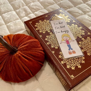 May include: A bookmark with a cartoon illustration of a girl with blonde hair and a pink shirt. The bookmark has the text "I'm hurt. I'm tired. I'm hungry." The bookmark is on top of a brown leather-bound book with gold accents.