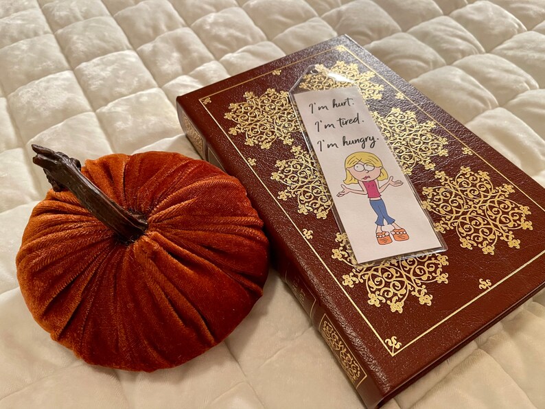 May include: A bookmark with a cartoon illustration of a girl with blonde hair and a pink shirt. The bookmark has the text "I'm hurt. I'm tired. I'm hungry." The bookmark is on top of a brown leather-bound book with gold accents.