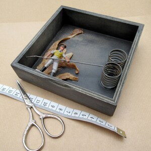 May include: A gray wooden box with a toy soldier inside. The soldier is holding a fishing rod and there are some dried leaves in the box. There are also three metal springs in the box.