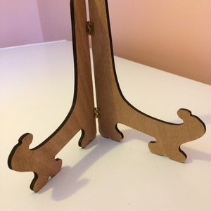 Può includere: Un cavalletto in legno con finitura naturale, caratterizzato da un design a cerniera per la regolazione. Il cavalletto ha una forma decorativa con gambe curve e una parte superiore leggermente curva. È progettato per contenere una cornice o un'opera d'arte.