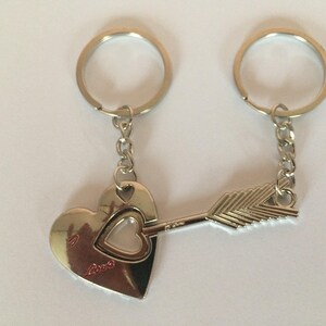 Könnte beinhalten: Silberne Schlüsselanhänger mit Herz- und Pfeildesign. Das Herz hat einen kleineren Herz-Ausschnitt und das Wort "love" eingraviert. Der Pfeil durchbohrt das Herz und symbolisiert die Liebe. Jeder Schlüsselanhänger hat einen silbernen Ring.