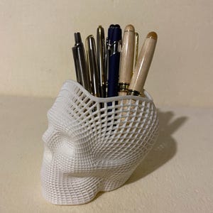 Peut inclure: Un porte-stylos blanc en forme de crâne imprimé en 3D rempli de divers stylos. Les stylos sont dans des tons de noir, d'or et de bois. Le support a un design en forme de treillis.