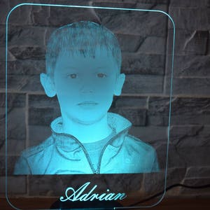 May include: A personalised LED night light with a portrait of a young person. The image is etched into a clear acrylic panel, lit with a soft blue light. The name "Adrian" is inscribed at the base of the light.