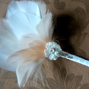 May include: A white and peach feather bouquet with a silver and pearl embellishment. The bouquet is attached to a white ribbon.