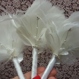 May include: Three white feather bouquets with silver wire and pearl accents. Each bouquet has a white satin ribbon handle and a small white lace embellishment.