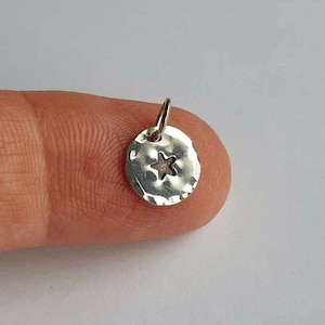 May include: A small, circular, silver-colored metal charm featuring a hammered, textured surface and a cut-out star shape in the center. The charm includes a small jump ring attached to the top, allowing it to be suspended from a chain or cord.