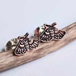 May include: A pair of copper-toned moth stud earrings featuring intricate black engraved details on the wings and body. Each earring has a metallic post back with a visible 925 stamp, indicating sterling silver construction for the backing.