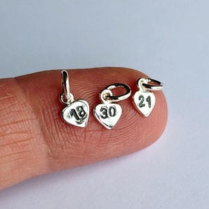 May include: Three small, silver heart-shaped charms. Each charm has a number: 18, 30, and 21. The charms are suspended from small loops, ready to be added to a necklace or bracelet. The background is a light, neutral colour.