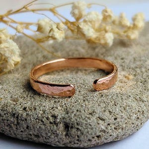 May include: A rose gold-toned open ring featuring a hammered, textured surface. The band is circular with a small gap at the front, showcasing a metallic finish that reflects light across its uneven, dimpled exterior surface.