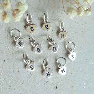 May include: A selection of silver heart-shaped charms, each bearing a different letter. The letters include S, E, H, R, G, D, B, K, M, and R. The charms are presented on a light grey surface.