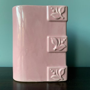 May include: A pink ceramic vase with a floral design. The vase has a rounded rectangular shape and three raised floral motifs on the front.