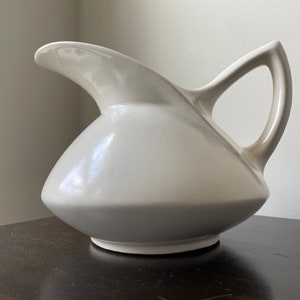 May include: A white ceramic pitcher with a unique, curved design and a handle. The pitcher has a smooth, glossy finish.