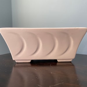 May include: A pale pink ceramic planter with a rectangular shape and a wavy design. The planter has three raised feet on the bottom.