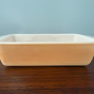 May include: A rectangular, light orange baking dish with a slightly flared rim. The dish is made of a solid, opaque material and sits on a dark brown wooden surface. The background is a solid light blue.