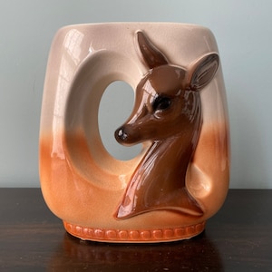 May include: A ceramic vase with a deer head design. The vase is a rectangular shape with a large, curved opening on one side. The deer head is brown, and the vase has a gradient of colors, from cream to orange.