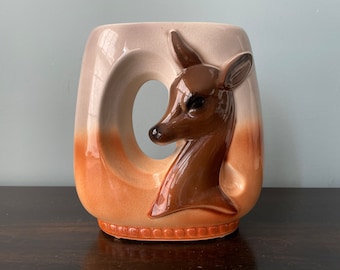 May include: A ceramic vase with a deer head design. The vase is a rectangular shape with a large, curved opening on one side. The deer head is brown, and the vase has a gradient of colors, from cream to orange.
