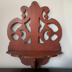 May include: A dark brown wooden wall shelf with intricate cutouts in the shape of swirls and leaves. The shelf has a scalloped edge and is mounted on a white wall.