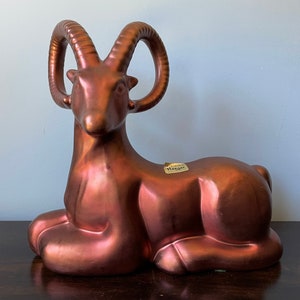 May include: A copper-colored ceramic sculpture of a sitting mountain goat with large curled horns. The sculpture has a label on the side that reads "Hanger".