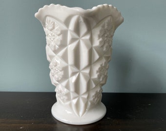 May include: A white milk glass vase with a snowflake pattern. The vase has a scalloped rim and a wide base.