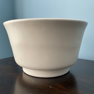 May include: A white ceramic bowl with a wide, rounded base and a slightly flared rim. The bowl is smooth and has a matte finish.