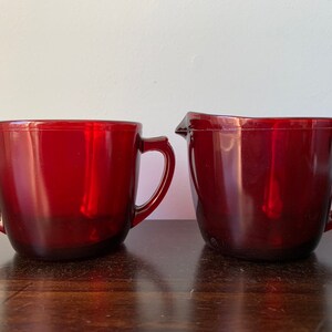 May include: A set of two red glass cups with handles. The cup on the left is smaller and the cup on the right is larger. Both cups are made of a clear glass with a red tint.